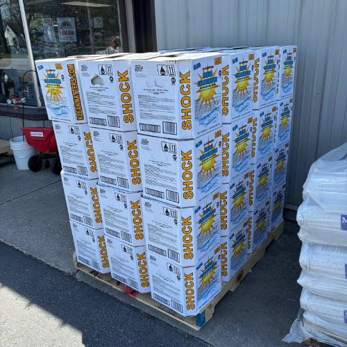 Pallet of pool shock boxes stacked on a sidewalk. Boxes display large yellow “SHOCK” text and a sun graphic. Nearby, other supplies are visible.