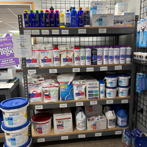 Shelves stocked with various pool care products, including bottles, bags, and buckets. Labels read 