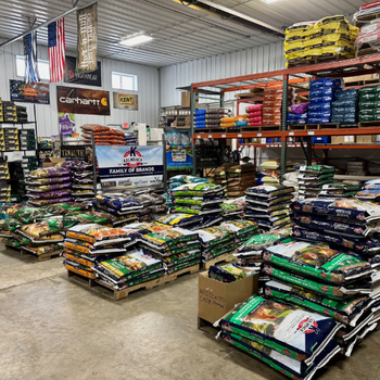 Farm supply store interior with neatly stacked pet food bags in various colors. Shelves display diverse brands; overhead banners and flags add a rustic touch.