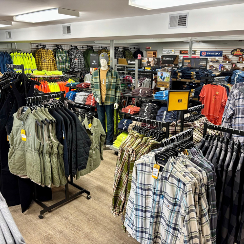 A clothing store interior features racks of plaid shirts, vests, and bright jackets. A mannequin dressed casually stands among neatly arranged apparel.