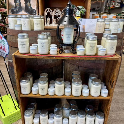 Wooden shelves display various scented candles in jars, with a decorative black lantern at the center. The setting evokes a cozy, inviting atmosphere.