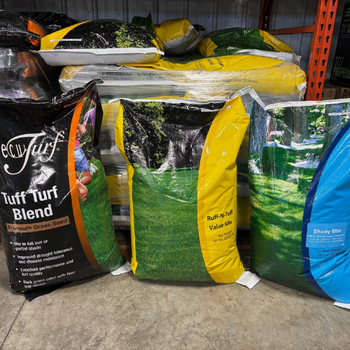 Bags of grass seed are displayed, featuring vibrant packaging labeled "Tuff Turf Blend," "Ruff-N-Turf Value Mix," and "Shady Mix" on a warehouse floor.