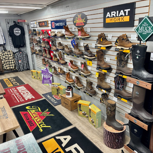 A store interior displays various work boots on wall shelves, featuring brands like Ariat and Twisted X. Mats with logos cover the wooden floor, creating a rugged, welcoming vibe.