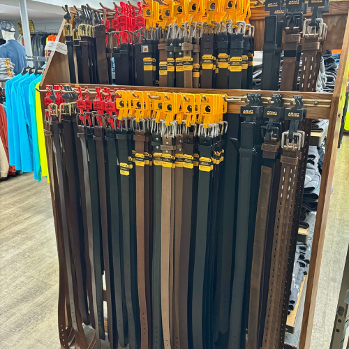 Display of various leather belts hanging on a wooden rack in a store. The belts are in black and brown shades with tags, surrounded by colorful shirts.
