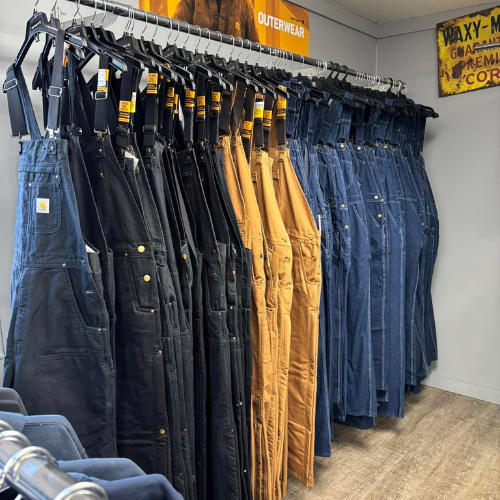 Racks of sturdy overalls in black, brown, and blue hang neatly in a store. The setting is tidy, with a rustic-style sign adding a vintage touch.