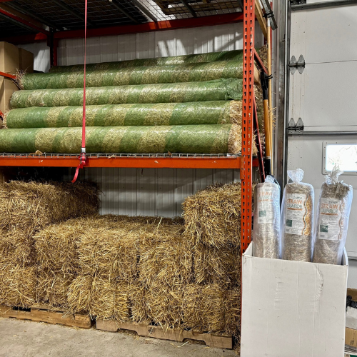 Shelves in a warehouse hold green rolled turf, stacked hay bales, and large bags of straw. The setting is organized, practical, and industrial.