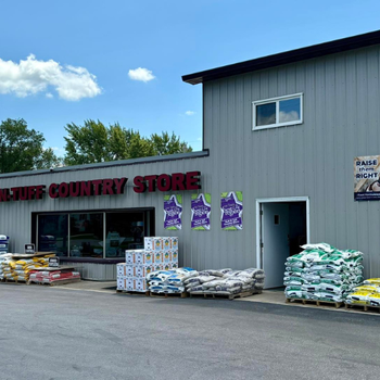 Exterior of a country store with a gray facade and red sign. Stacks of feed bags and supplies are displayed outside. The atmosphere is rural and inviting.