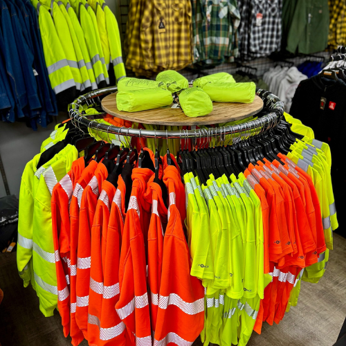 A circular display of high-visibility workwear with bright orange and yellow shirts with reflective stripes. Surrounding shelves hold folded jackets.