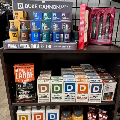 Duke Cannon display with colognes, lip balm, and soap bars. Shelf messages read 