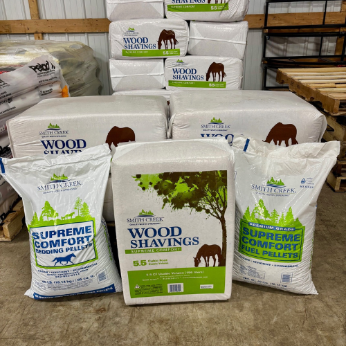 Bags of wood shavings and bedding pellets are stacked in a warehouse. The packaging features green and white colors with tree and horse imagery.